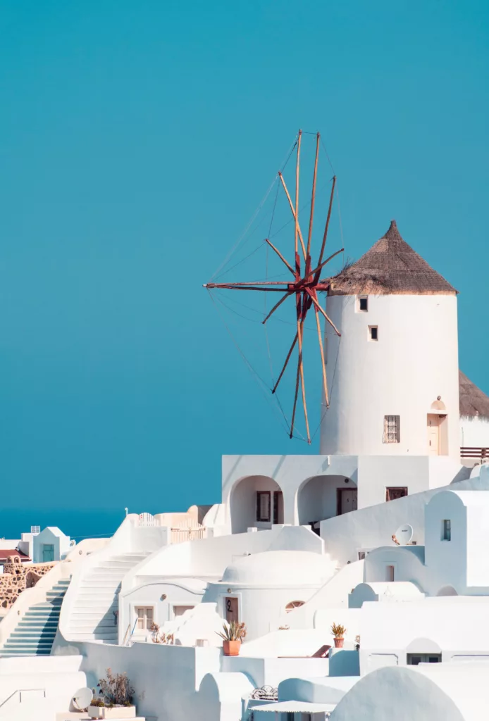 A picturesque view of Mykonos featuring a classic windmill amid a cluster of white-washed buildings against a clear blue sky.