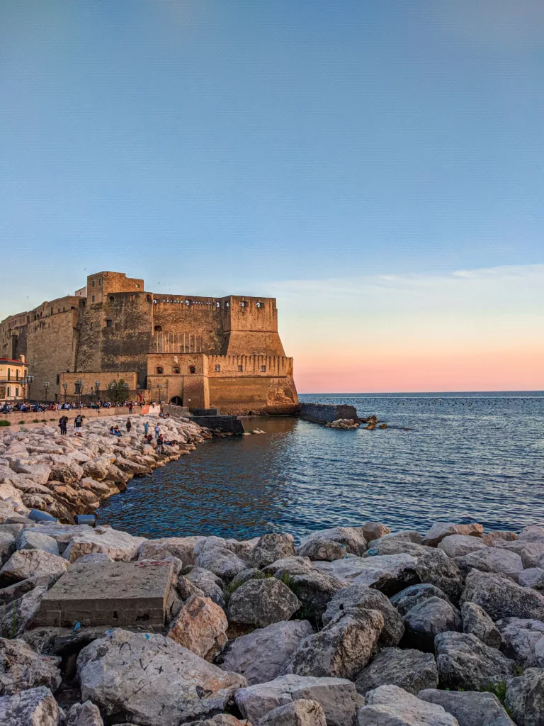 A view of Castel dell'Ovo, a historic seaside fortress in Naples, Italy. The castle sits on a rocky promontory surrounded by the sea, with a warm glow from the setting sun reflecting on its ancient walls.
